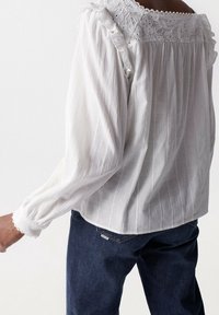 Weißes Blusenoberteil mit vertikalen Streifen, Rüschenkragen, Spitzendetails und langen Ärmeln, kombiniert mit dunkelblauen Jeans.