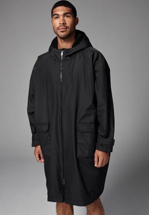 Schwarzer wasserdichter Mantel mit Reißverschluss, übergroßer Schnitt, großen Taschen und einer weichen, gefütterten Kapuze. Glatte Außenfläche, leichtes Design.
