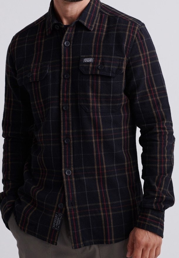 Mand iført en sort, ternet, langærmet skjorte med knapper og brystlommer, stående med den ene hånd i lommen mod en ensfarvet baggrund.