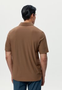 Brūns polo krekls ar mīkstu tekstūru, īsām piedurknēm un apkakli. Satur smalku logo izdrukātu uz augšējās muguras.