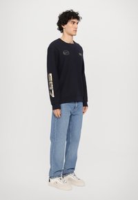 Jūras spēku džemperis ar zelta akcentiem uz piedurknēm un logo detaļām, brīvs piegriezums, apaļa kakla izgriezums. Apvienots ar gaišu džinsa audumu biksēm un baltām kedām.