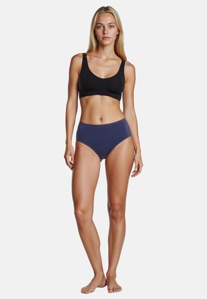 Young woman standing barefoot wearing a black sports bra and navy high-waisted underwear against a plain white background.