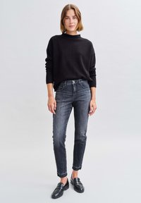 Maglione nero lavorato a maglia con colletto alto, abbinato a jeans skinny grigi scuri e mocassini neri. Il maglione ha una vestibilità rilassata e polsini a costine.