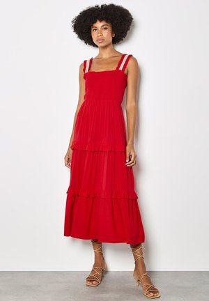 Kobieta z kręconymi włosami w czerwonej, wielowarstwowej sukience midi bez rękawów z falbanami i beżowych sandałkach na obcasie, stoi przy białej ścianie.