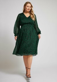 Grön prickig klänning med v-ringning, långa genomskinliga ärmar och elastisk midja. Flödande kjol som slutar ovanför vristen, tillsammans med svarta remsandaler.