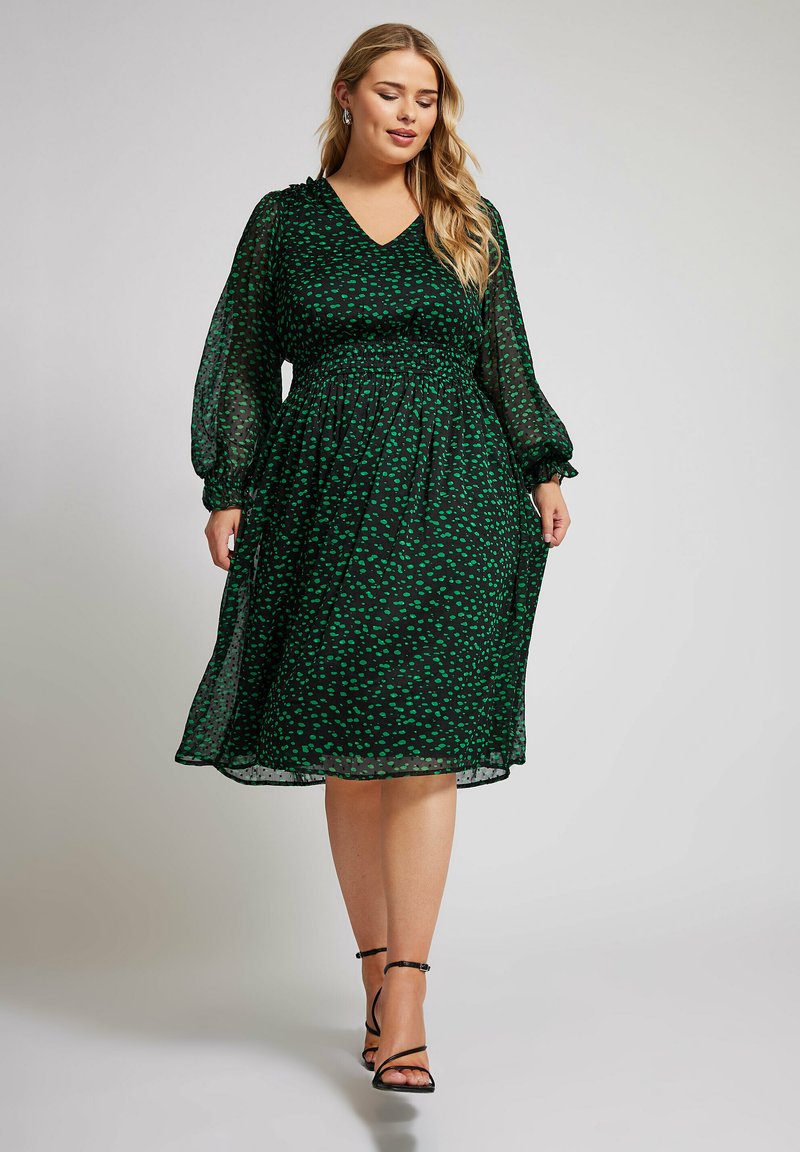 Grön prickig klänning med v-ringning, långa genomskinliga ärmar och elastisk midja. Flödande kjol som slutar ovanför vristen, tillsammans med svarta remsandaler.