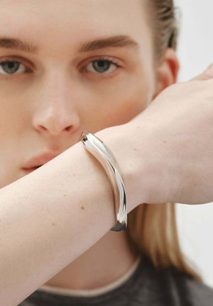 Pulsera de metal plateado con un diseño curvado, textura suave y superficie pulida, llevada en una muñeca de piel clara.