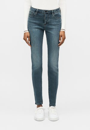 Kobieta ubrana w niebieskie jeansy o średnim stanie i dopasowanym kroju, biały długi rękaw oraz białe trampki, stojąca na tle jasnego, jednolitego tła.