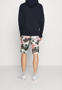 Mężczyzna w granatowej bluzie z kapturem, białych szortach w tropikalny wzór, szarych skarpetkach i białych trampkach stoi twarzą do białej ściany.