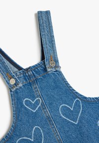 Denimoveraller med justerbara axelremmar, en blåtvätt och vita hjärtan. Metallaccent och kontrastsömnadsdetaljer.