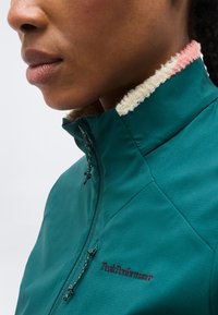 Κοντινό πλάνο ενός ατόμου που φοράει ένα τζάκετ Peak Performance σε τιρκουάζ χρώμα με φερμουάρ και γιακά επενδυμένο με λευκή και ροζ μάλλινη επένδυση.