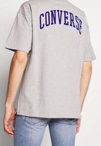 Hombre vistiendo una camiseta gris con "CONVERSE" en letras moradas en la espalda, combinada con jeans de mezclilla azul claro.