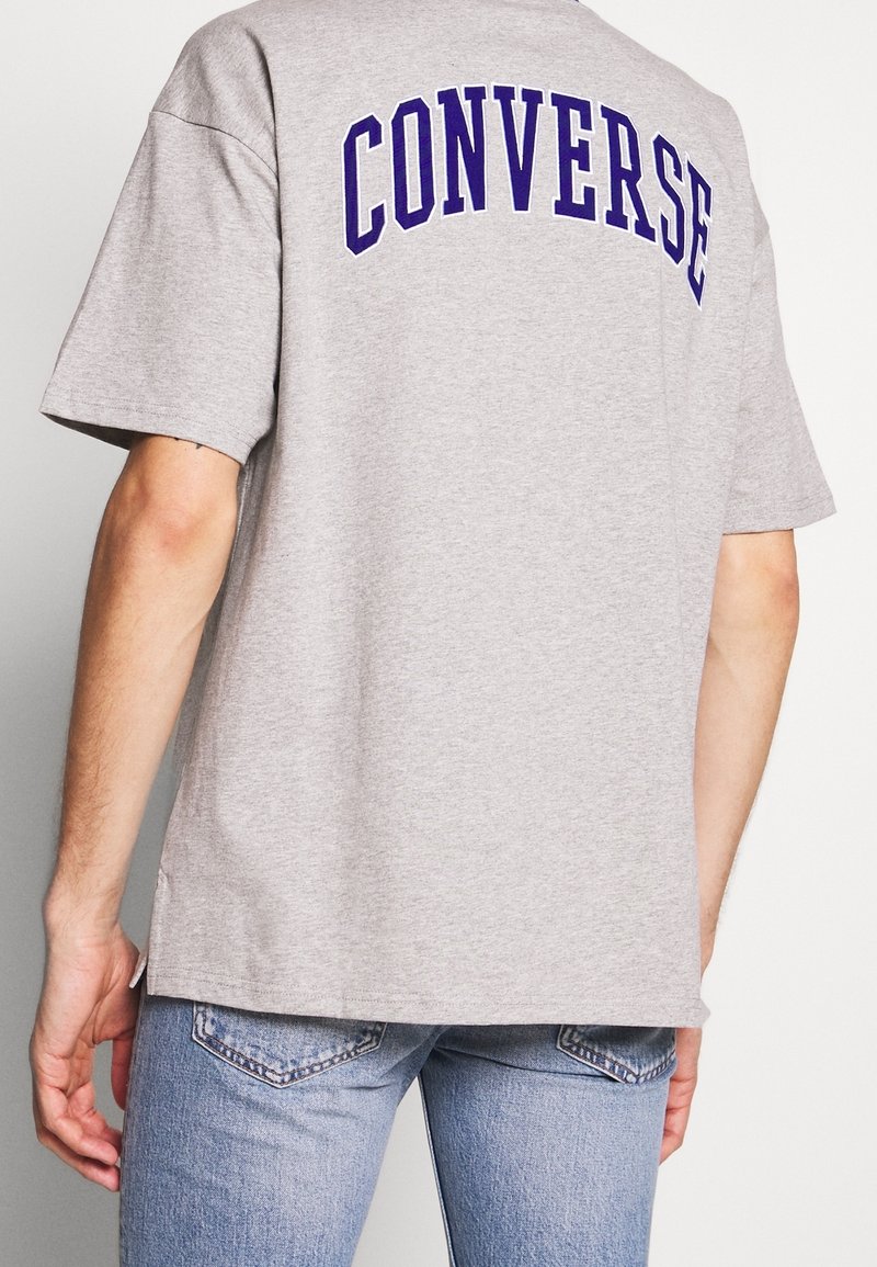 Hombre vistiendo una camiseta gris con "CONVERSE" en letras moradas en la espalda, combinada con jeans de mezclilla azul claro.