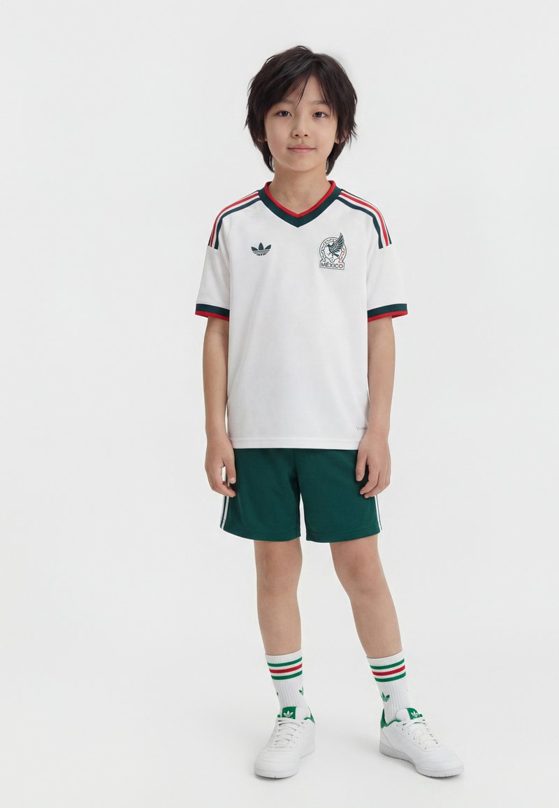 Νεαρός αγόρι, όρθιος, φοράει λευκή φανέλα ποδοσφαίρου του Μεξικού με πράσινο σορτς, ριγέ κάλτσες και λευκά αθλητικά παπούτσια σε απλό φόντο.