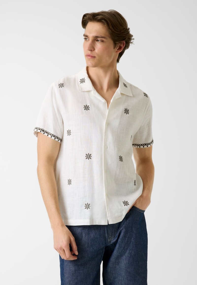 Jeune homme portant une chemise blanche à manches courtes avec des motifs noirs brodés et un jean bleu foncé, regardant sur le côté.