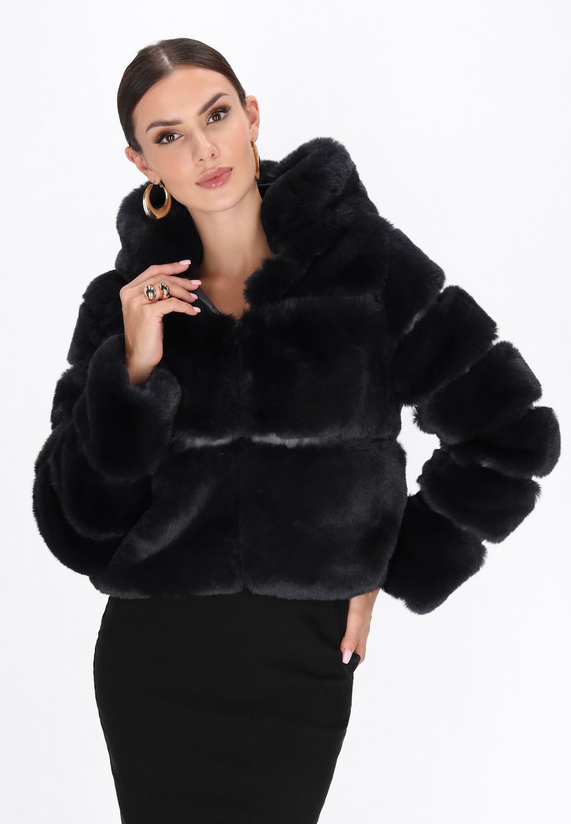 Sort faux fur cropped jakke med hætte, der har en blød tekstur og brede ærmer. Parret med en tætsiddende sort nederdel. Guld hoops øreringe og ring.