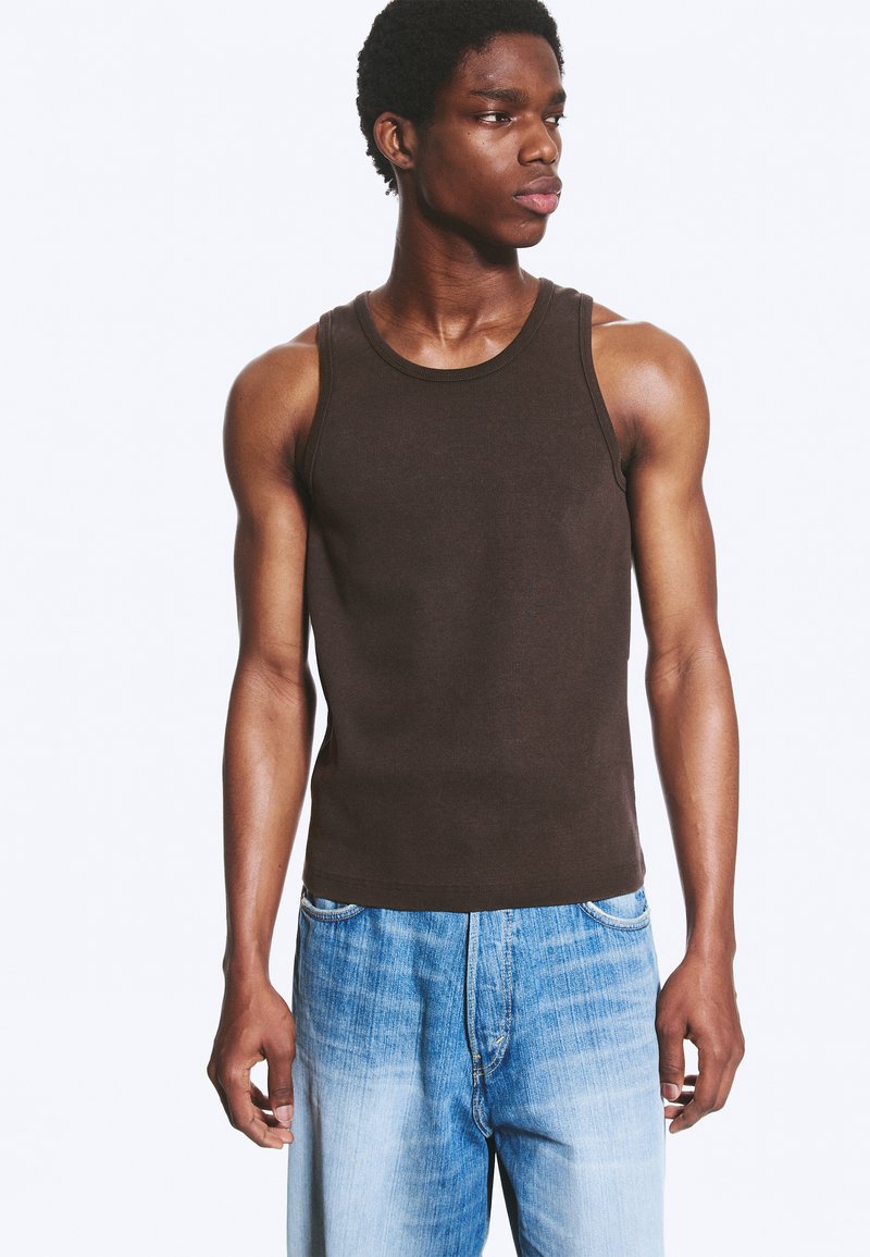 Jeune homme portant un débardeur marron foncé et un jean bleu clair, regardant vers sa gauche sur un fond clair uni.