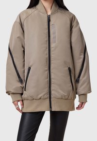 Beige Bomberjacke mit durchgehendem Reißverschluss, gerippten Bündchen und zwei Seitentaschen. Verfügt über schwarze Reißverschlussdetails und einen kleinen Aufnäher auf der Brust.