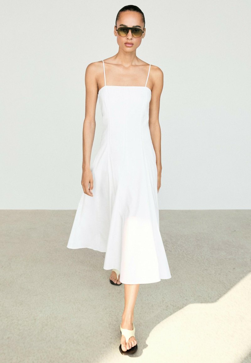 Woman wearing a sleeveless white midi dress, white flip-flop sandals, and olive green sunglasses walking on a light floor.