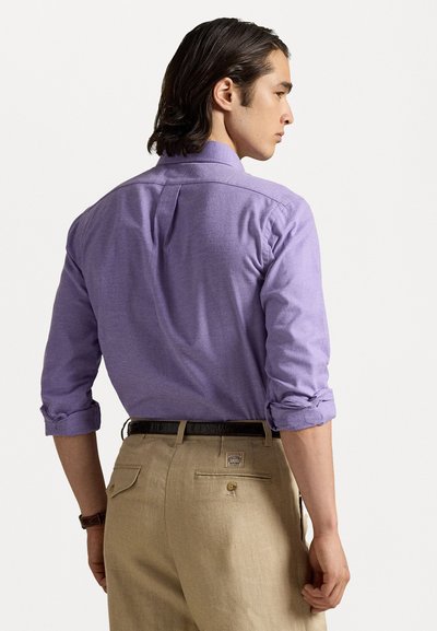 Cămașă lungă cu mânecă, de culoare violet deschis, cu mansete răsucite și pliu pe spate, asortată cu pantaloni bej care au un buzunar la spate și un logo cusut.