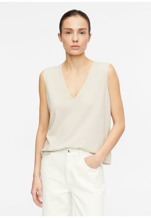 Kobieta o brązowych włosach w bezrękawnym beżowym topie z dekoltem w serek i białych spodniach, stojąca z jedną ręką w kieszeni na jednolitym tle.