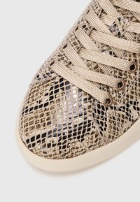 Sneaker con motivo in pitone beige e nero, lacci beige infilati attraverso sei occhielli in metallo e suola beige su sfondo bianco.