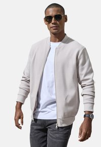 Helle beige Bomberjacke aus strukturiertem Stoff mit gerippten Bündchen und Saum, getragen über einem weißen T-Shirt, kombiniert mit grauen Jeans.