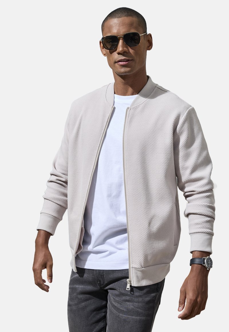 Helle beige Bomberjacke aus strukturiertem Stoff mit gerippten Bündchen und Saum, getragen über einem weißen T-Shirt, kombiniert mit grauen Jeans.