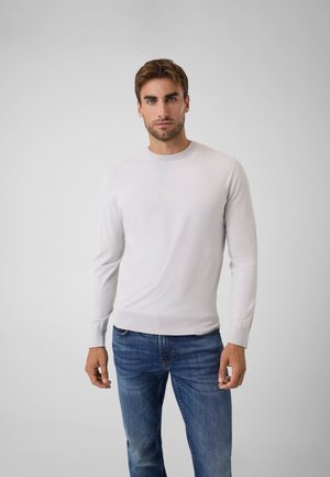Homme aux cheveux courts bruns portant une chemise unie gris clair à manches longues et un jean bleu, debout devant un fond blanc.