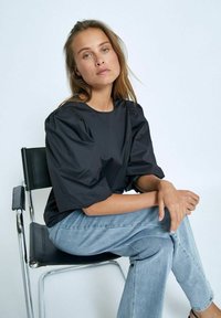 Blusa nera strutturata con maniche a sbuffo, abbinata a jeans di denim blu chiaro. Modello seduto su una sedia di cromo contro uno sfondo neutro.