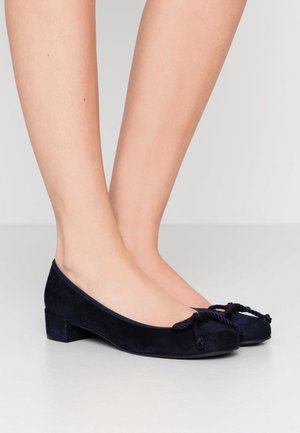 Pies que llevan bailarinas bajas de ante azul marino con detalles de moño de cuerda trenzada sobre un suelo gris claro.