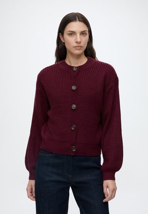 Kvinde iført en mørk lilla strikket cardigan med store knapper og mørkeblå jeans, stående mod en ensfarvet hvid baggrund.