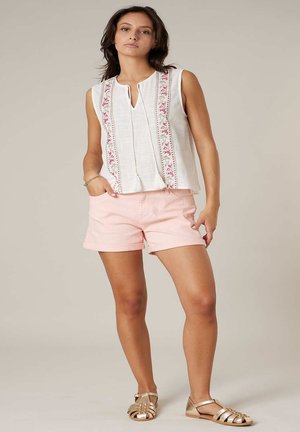 Femme portant un haut blanc sans manches avec broderie florale, un short rose et des sandales dorées, debout avec une main dans la poche.