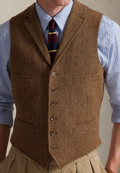 Καφέ γιλέκο με ψαροκόκκαλο, πέντε κουμπιά, εγκοπές στους πέτους και τσέπη στο αριστερό μέρος του στήθους, φορεμένο πάνω από μπλε ριγέ πουκάμισο και γραβάτα με σχέδια.