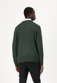 Grüner Strickpullover mit hohem Kragen und geripptem Saum, der eine strukturierte Oberfläche aufweist. Wird über dunklen Hosen getragen und zeigt eine lockere Passform.