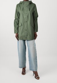 Grön vattentät jacka med huva, tryckknappar, två framfickor och dragsnören, kombinerad med ljusblå vida jeans och klackar.