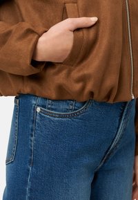 Hnědá semišová bomber bunda se zipem vpředu a bočními kapsami, doplněná modrými džínami s klasickým pětikapsovým designem.