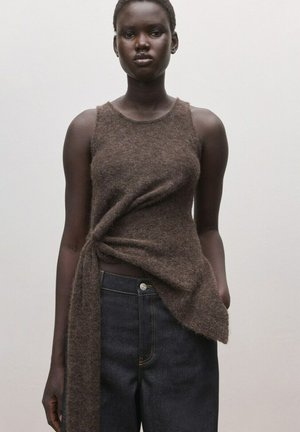 Mujer de piel oscura que lleva una blusa marrón sin mangas y peluda con un detalle de nudo asimétrico y vaqueros de mezclilla oscuros, sobre un fondo sencillo.