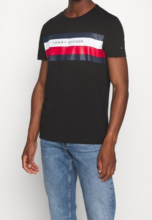 Mann trägt ein schwarzes Tommy Hilfiger T-Shirt mit roten, weißen und Marineblauen Streifen und blaue Jeans vor einfarbigem Hintergrund.