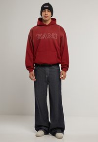 Crvena dukserica s kapuljačom i "KANI" logom, u kombinaciji s širokim tamnim trapericama i bijelim tenisicama. Crna pletena kapa na glavi.