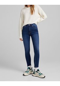 Vysoké modré džíny slim fit, v kombinaci s bílým pleteným svetrem a robustními teniskami s zelenými akcenty a bílou podrážkou.