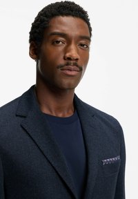 Granatowa marynarka w jodełkę z wyciętymi klapami, teksturowaną tkaniną i poszetką z wzorzystym akcentem. Noszona na ciemnym T-shircie.