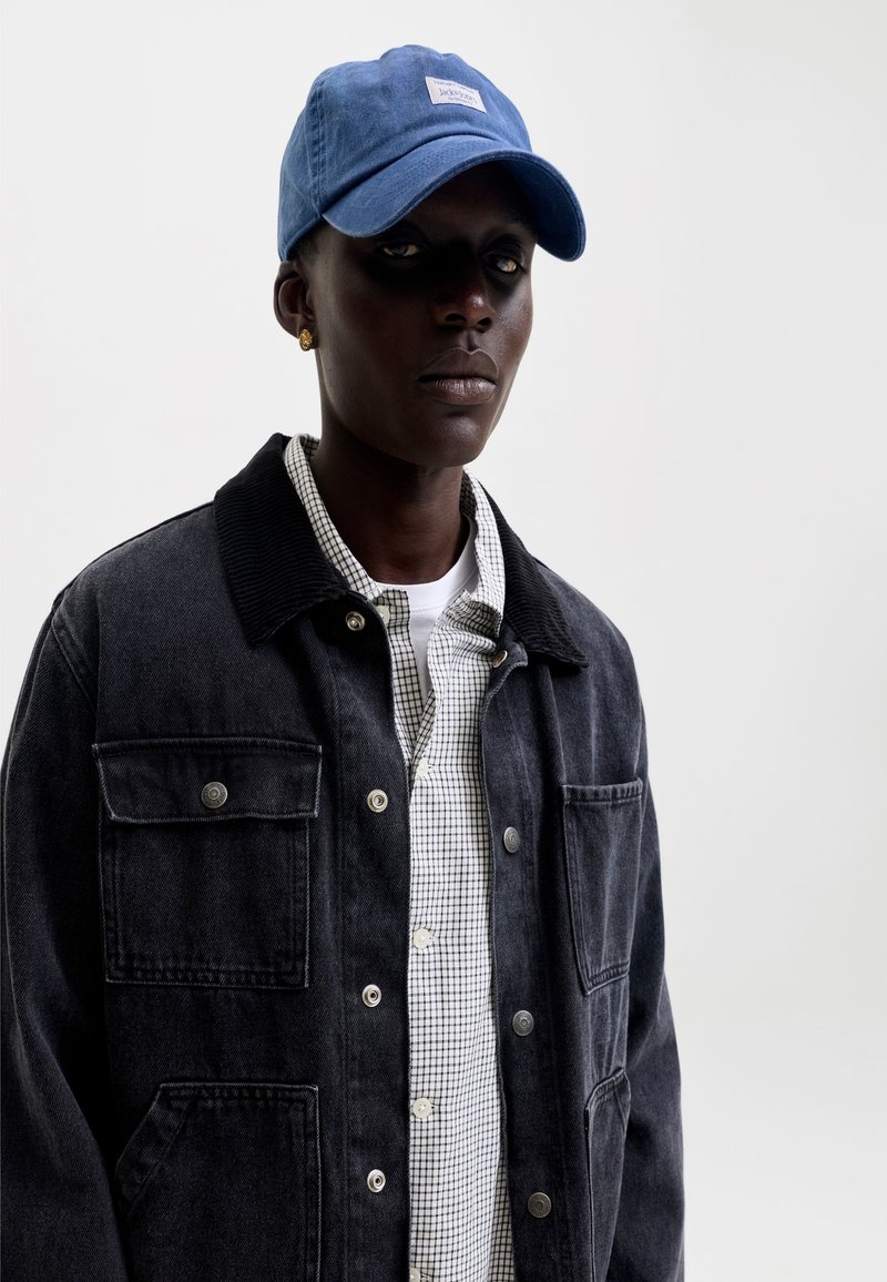 Personne à la peau foncée portant une casquette bleue, des boucles d'oreilles dorées, une veste en jean noire et une chemise blanche à carreaux sur un fond clair uni.