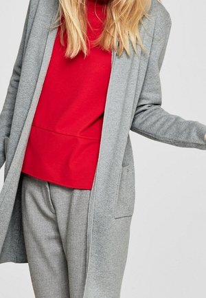 Cardigan long gris avec poches sur un top rouge, associé à un pantalon gris clair. Tissu doux, coupe décontractée, sans fermetures visibles.