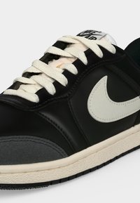 Μαύρο και γκρι αθλητικό παπούτσι Nike με λευκά κορδόνια, λευκό λογότυπο Nike και λευκή σόλα με μαύρο πέλμα.