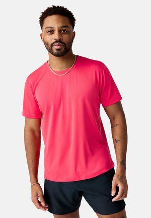 Mann mit Bart und Tätowierungen, der ein leuchtend pinkes kurzärmliges Hemd und schwarze Shorts trägt und vor einem schlichten weißen Hintergrund steht.