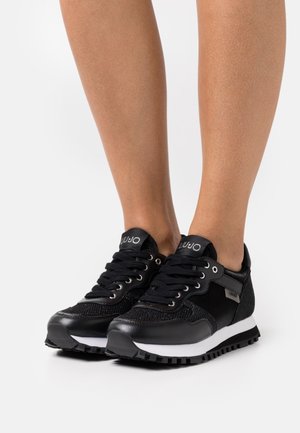 Baskets noires avec semelles blanches et détails en mesh portées pieds nus, avec une étiquette de marque sur la languette et le côté de chaque chaussure.