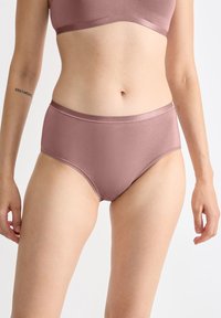 Vrouw in mauve naadloze ondergoed en bijpassende beha, met een tatoeage op haar linker binnenarm, staand tegen een plain witte achtergrond.