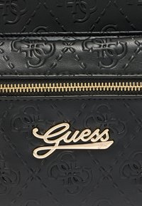 Μαύρο δερματίνη με υφή και ανάγλυφα σχέδια, διακοσμημένο με έντονο χρυσό λογότυπο "Guess" και χρυσό φερμουάρ.