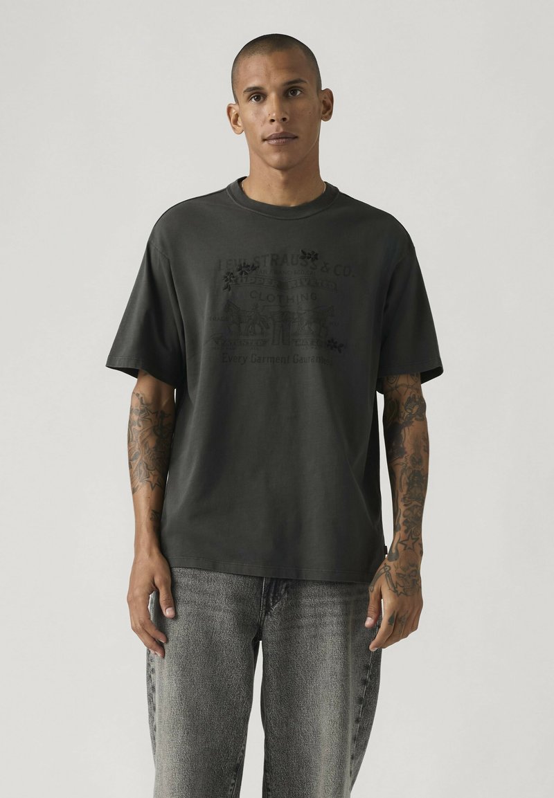 Homme aux cheveux courts et tatouages sur les deux bras, portant un T-shirt graphique sombre et surdimensionné ainsi qu'un jean gris clair, debout devant un fond uni.